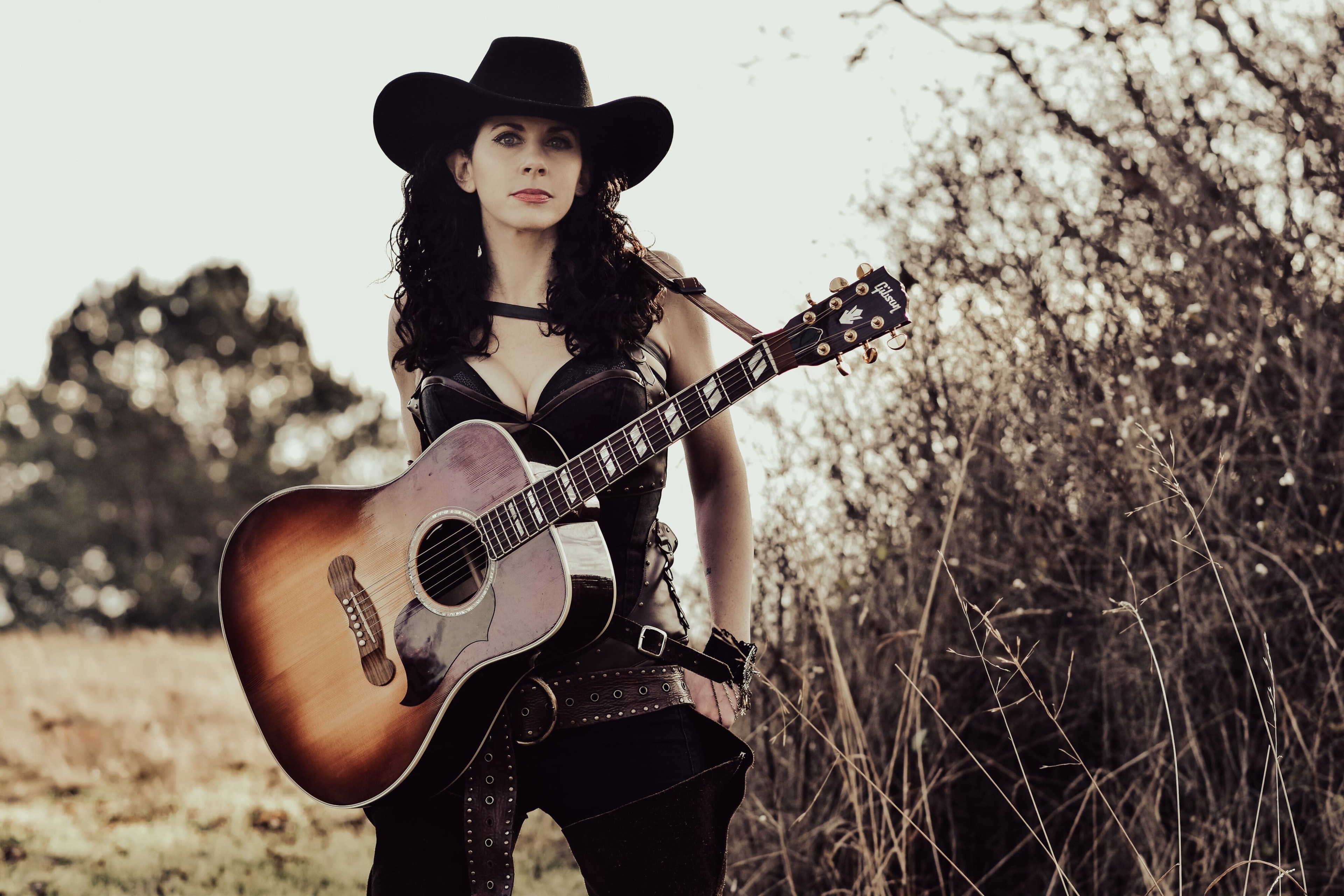 Melissa Endean with guitar slung over shoulder in dramatic leather outfit against a blue sky